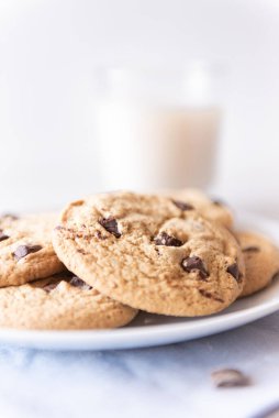 Beyaz tabakta çikolatalı kurabiyeler, beyaz masada bir bardak süt ve mavi peçete.