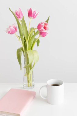 Cam bir vazoda pembe laleler, beyaz bir masada kupalar ve kitaplar..