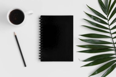 Çalışma alanı aksesuarları ve beyaz bir masanın üzerinde bir palmiye yaprağı olan siyah bir defterin üst görünümü.