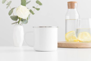 Enamel kupası vazoda çiçekler, cam ve beyaz bir masada bir şişe..