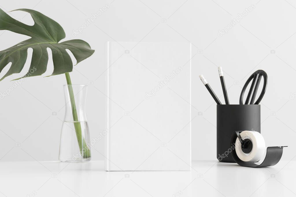 White book mockup with a monstera leaf in a glass vase and workspace accessories on a white table.