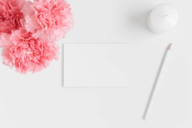 Beyaz bir kartın üst görüntüsü. Üzerinde pembe karanfiller ve çalışma alanı aksesuarları olan beyaz bir masa..