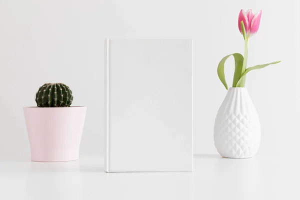 White book mockup with a pink tulip and a cactus in a pot on white table.