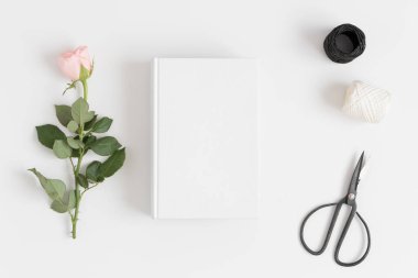 Beyaz bir masanın üstünde pembe bir gül, makas ve sicimle beyaz bir kitabın üst görüntüsü..
