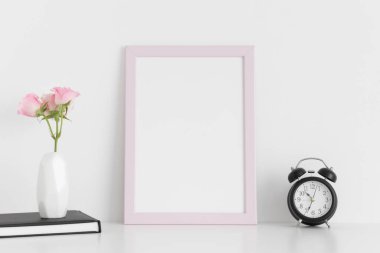 Üzerinde çalışma alanı aksesuarları ve pembe güller olan pembe çerçeve modeli beyaz bir masa üzerinde bir vazoda..