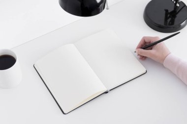 Bir kupayla defter modelini aç, beyaz bir masada lamba ve not alan bir kadın..