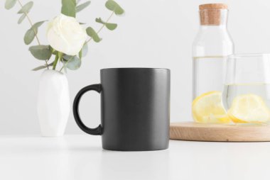 Siyah kupa, vazoda çiçekler, cam ve beyaz masada bir şişe..