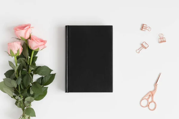 Top view of a black book mockup with a bouquet of pink roses and workspace accessories on a white table.
