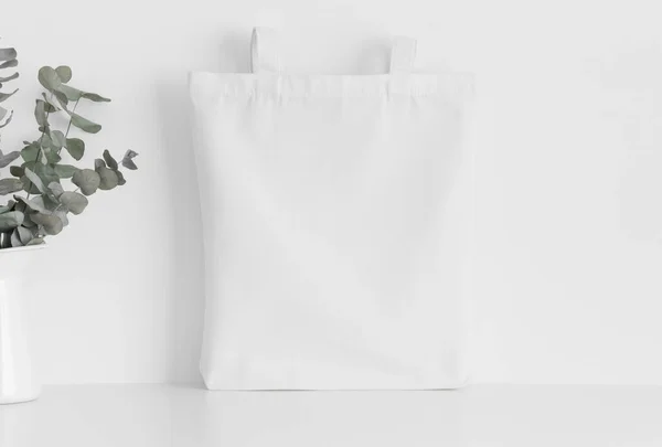 White tote bag mockup with a eucalyptus in a vase on a white table.