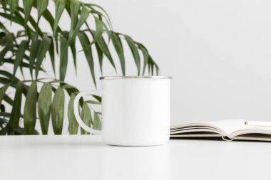 Beyaz bir masa ve bir palmiye yaprağı üzerinde not defteri olan Enamel kupası..