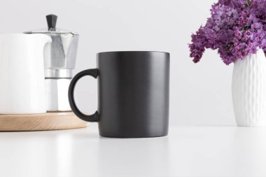 Siyah kupa moka kabı ve beyaz masadaki vazoda bir demet leylak..