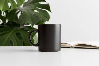 Siyah kupa, beyaz bir masanın üzerinde bir defter ve bir canavar bitkisi..