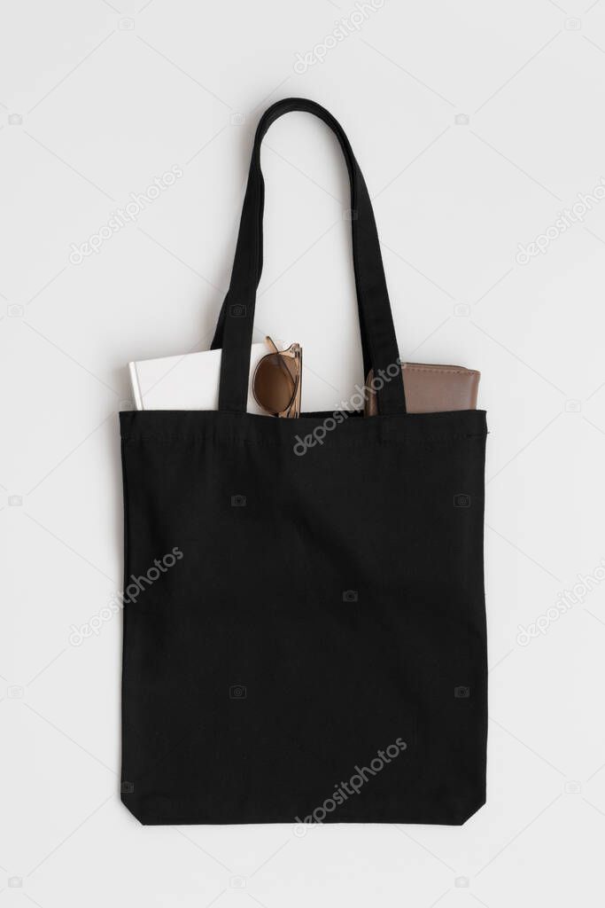 Black tote bag mockup with a book, wallet and glasses on a white table.