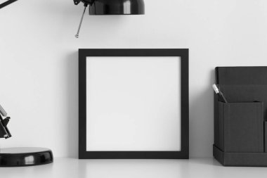 Beyaz masa üzerinde lamba ve çalışma alanı aksesuarları olan siyah kare çerçeve modeli.