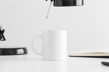 Beyaz kupa bir lamba ve beyaz bir masanın üzerinde bir defter..