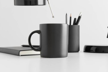 Beyaz bir masada lamba, defter ve çalışma alanı aksesuarlarıyla siyah kupa modeli..