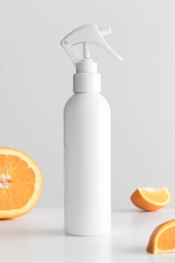 Beyaz kozmetik tetik püskürtücü şişesi beyaz bir masada portakallarla modellenmiş..