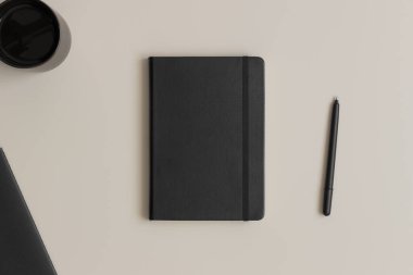 Siyah defter modeli, bej bir masada bir fincan kahve ve çalışma alanı aksesuarları..