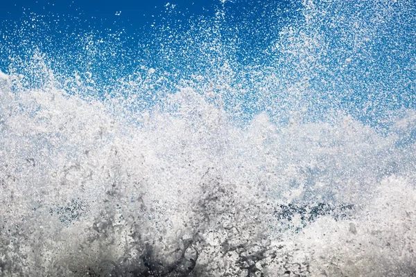 Spray from waves crashing over a rock, with a blue sky behind