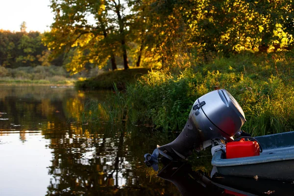 Part of metal blue boat with large powerful gray gasoline engine and beautiful red fuel canister, floating in water near shore against background of green grass and trees on sunny autumn evening — Stock Image Part Metal Blue Boat Large Powerful Gray Gasoline Engine Beautiful Royalty Free Stock Photos