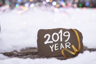 Brown piece of cardboard with the inscription 2019 YEAR on a background of snow and bright lights, close-up