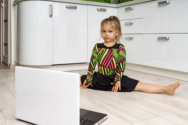 little girl 4 years old in a gymnastic leotard is engaged in online gymnastics at home and looks at the laptop. sitting on a twine
