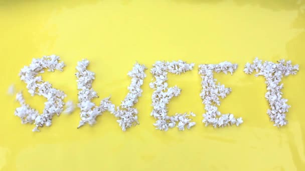 De inscriptie Sweet is geschreven in witte bloemen van lila op een gele achtergrond. Het woord Sweet is geschreven met lila bloemen. Bloeminscriptie Zoet. — Stockvideo