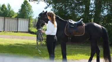 Beyaz gömlekli ve siyah pantolonlu bir kız ve güneşli yaz eğitiminde siyah bir at.