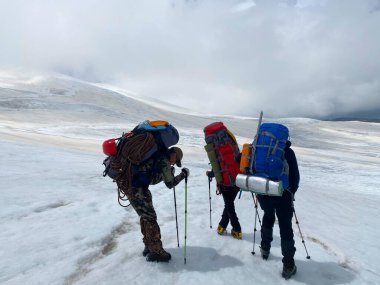 Sırt çantaları ve yürüyüş direkleri olan üç yorgun dağcı Elbrus 'un yamaçlarında duruyor. Kuzey Elbrus bölgesinin karlı yamaçları. Dağ kış manzarası.
