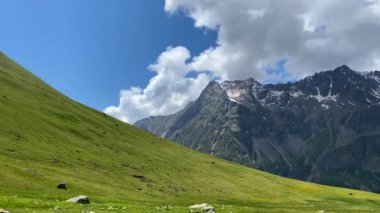 Kuzey Elbrus bölgesinin yeşil kayalık yamaçları. Güzel bir dağ manzarasının havadan görünüşü. Beyaz bulutlu mavi gökyüzü. Uzakta karla kaplı zirveler.