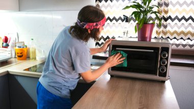 Temizlikçi mikrodalga fırını temizler. Garson kadın mutfağı temizliyor. Bir kadın ev işleri yapar. Bir eşin görevleri. Mavi bir tişört ve pembe bir bandana giyiyor..