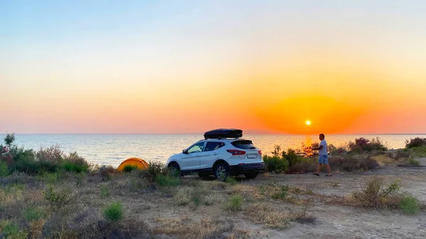 Tudakul rezervuarında kamp yapmak. Özbekistan 'da arabayla seyahat etmek. Deniz manzaralı çarpıcı turuncu gün batımı. Okyanusun yanındaki plajda beyaz bir araba park halinde.