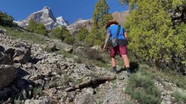 Alaudinsky göllerine yolculuk. Kovboy şapkalı bir adam yeşil ağaçların ve dağlık tepelerin ardında kayalık bir dağ yolunda yürüyor. Seyahat ve macera.