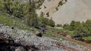 Bir adam dağ nehri boyunca kayalık bir tepeye tırmanır. Kovboy şapkalı yalnız bir turist Tacikistan 'ın dağlık bir bölgesinde yürüyor. Alaudinsky göllerine yolculuk.