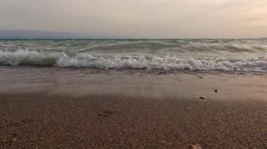 Dalgalar kumlu bir sahile yuvarlanırken ve köpük oluştururken aşağıdan bir manzara. Gökyüzü bulutlarla kaplıdır ve güneş görünür değildir. Denizde tatil. Göl kenarında tatil.