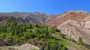Sert, çorak bir dağ manzarasının ortasında ağaçlarla kaplı küçük yeşil bir vadinin havadan görünüşü. Önünde yemyeşil bir orman olan bir dağ sırası..