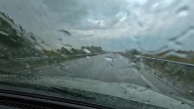 Yağmurlu bir yolda, ön cam silecekleri açık, kapalı bir şekilde bir araba sürüyor. Arabayla yolculuk. Sahne biraz kasvetli. Kötü hava