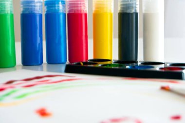 İçinde akrilik boya konteynerleri ve içinde su ve fırça bulunan plastik bir bardak olan renkli boyalardan yapılmış bir çocuk resmi olan beyaz kağıt.