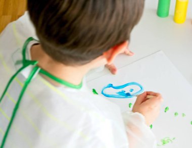 Bir fırça ve mavi boyayla resim yapan küçük bir çocuk ve beyaz bir masa üzerinde beyaz bir kağıt ve renkli boyalı kaplar.