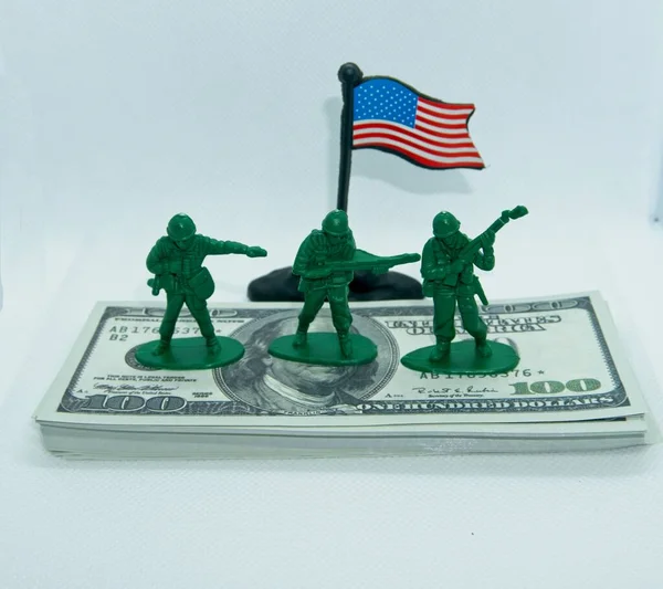 Plastic figures of miniature soldiers in gray and green colors on top of a wad of hundred dollar bills with a U.S. flag on the side with a white background