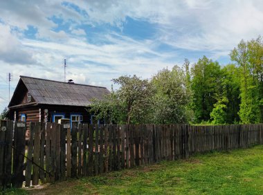 Yeşil bir ormanın yakınındaki ahşap bir çitin arkasındaki eski köy evi bulutlu mavi bir gökyüzüne karşı.