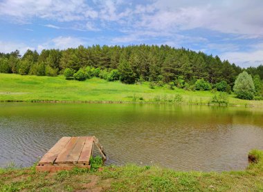 Yüzen bir göl üzerinde küçük ahşap bir köprü diğer taraftaki yeşil ormanın arka planında ve bulutlu güzel mavi bir gökyüzü.