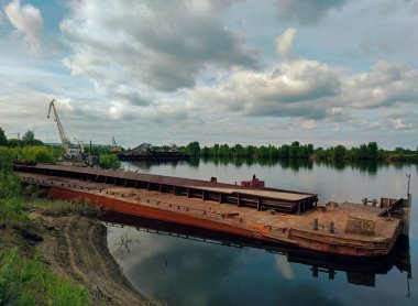 Güzel mavi bulutlu bir gökyüzünün arka planında rıhtımın yanındaki nehir kıyısında terk edilmiş eski paslı, kendi kendine hareket etmeyen mavna.