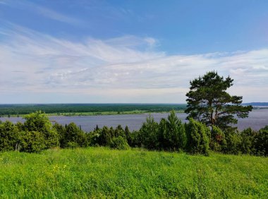 Bir dağın tepesinde yeşil çimen ve ağaçlarla dolu çayır bir nehrin arka planına ve güneşli bir günde bulutlu harika mavi bir gökyüzüne karşı.