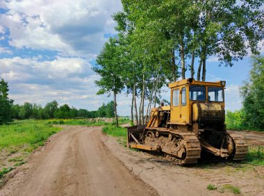 Yaşlı sarı buldozer, ağaçların yanındaki kır yolunun kenarında güneşli bir günde bulutlu mavi bir gökyüzüne karşı duruyor.