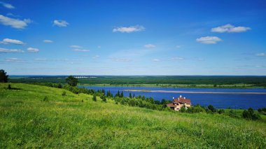 Nehir kıyısında yeşil bir dağ yamacındaki ev güneşli bir günde bulutlu mavi gökyüzüne karşı.
