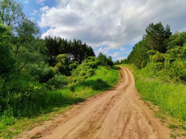 Güneşli mavi gökyüzü, yeşil çimenler ve ağaçlar arasında yokuş yukarı giden bir kır yolu üzerindeki bulutlarla.