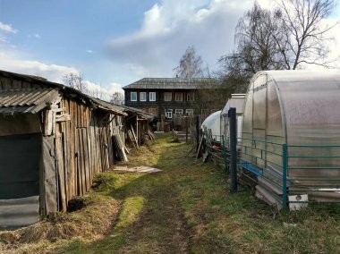 Mavi gökyüzüne karşı harap olmuş ahşap bir evin yanında seraları ve garajları olan eski bir avlu.