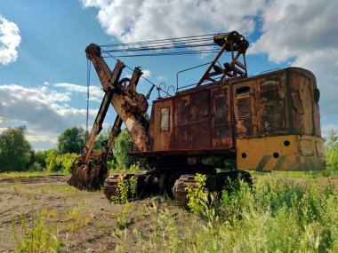Güneşli bir günde mavi bulutlu bir gökyüzüne karşı yeşil çimlerle kaplı kumlu bir platformda paslı büyük bir kazıcı.