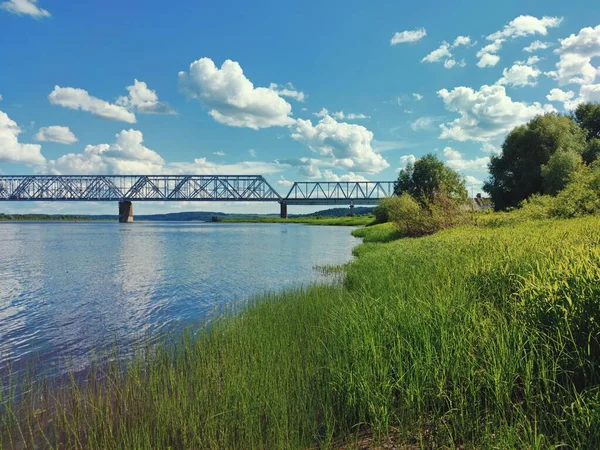 green river bank in front of a bridge against a blue sky with clouds on a sunny day